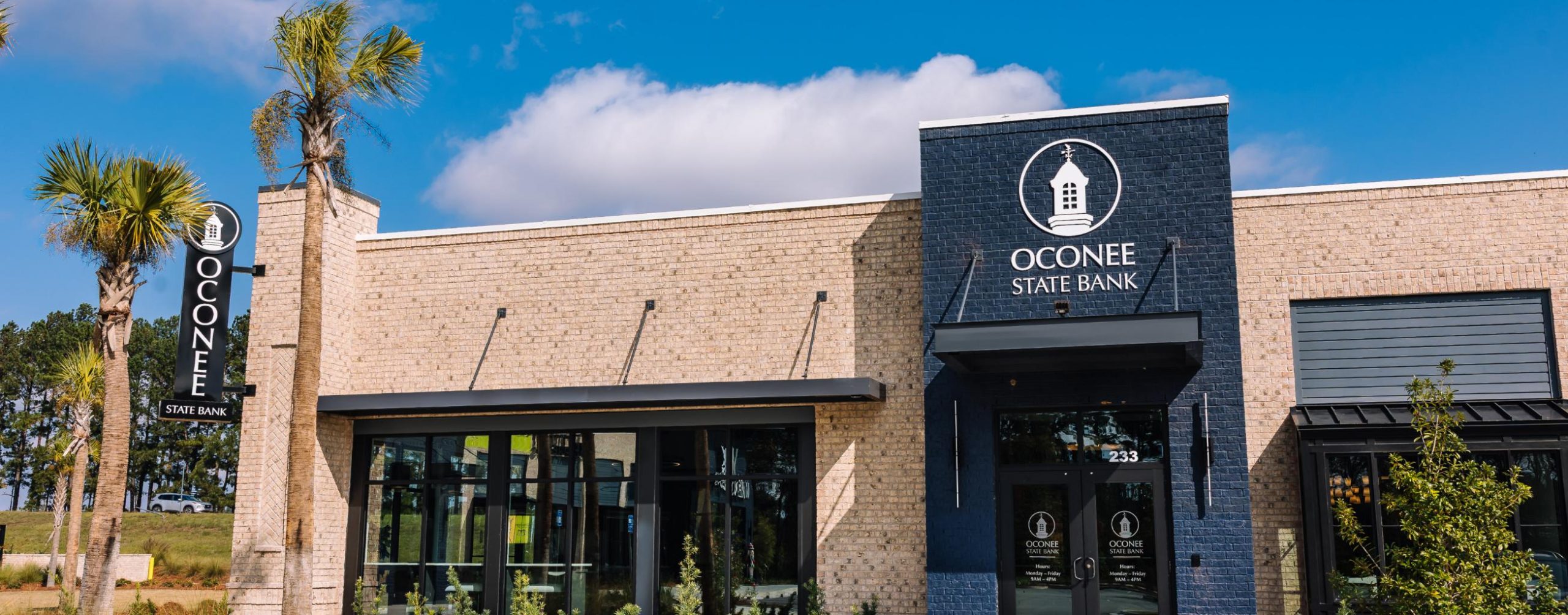 A modern Oconee State Bank building with light brick walls, large windows, and a dark blue sign featuring the bank’s logo. Tall palm trees and a clear sky with some clouds are visible outside.