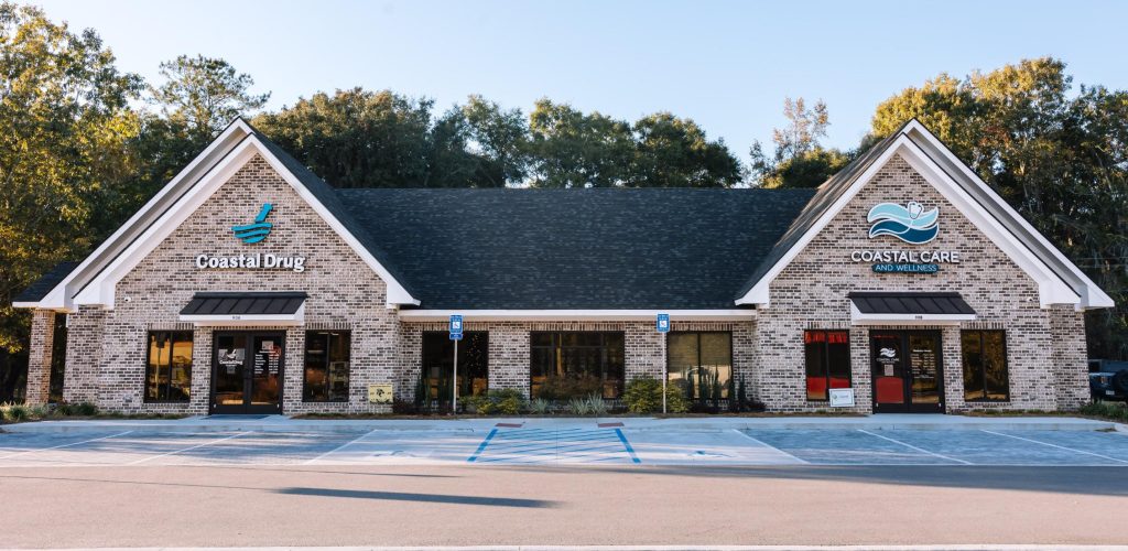 A brick building with two businesses: Coastal Drug on the left and Coastal Care on the right. Both have large front windows, signs with blue logos, and an accessible parking space in front. Trees are visible in the background.