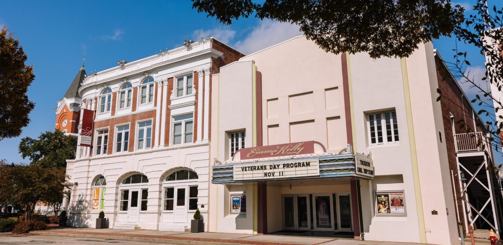 A historic brick and white theater building with a marquee is seen on a sunny day, with trees framing the scene and posters displayed near the entrance.