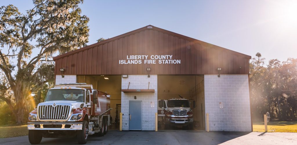 A brick fire station with Liberty County Islands Fire Station on the front. Two fire trucks are parked, one outside and one inside the open garage bays, with trees and sunlight in the background.