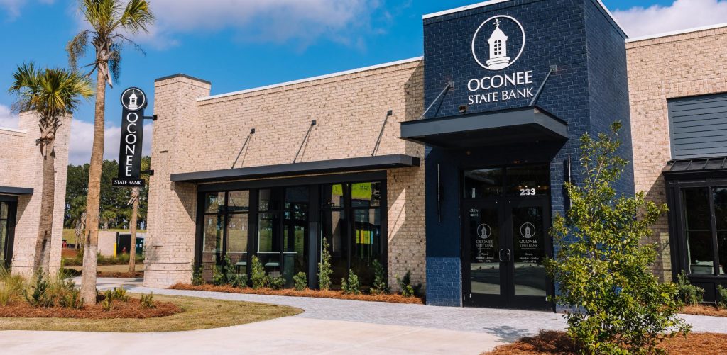 A modern brick building with a navy blue entrance displays the Oconee State Bank logo and name. Palm trees and landscaping are visible in front, with a clear blue sky overhead.