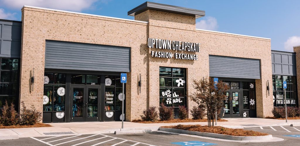 A brick storefront for Uptown Cheapskate Fashion Exchange with large windows, visible signage, and empty accessible parking spaces in front under a blue sky.