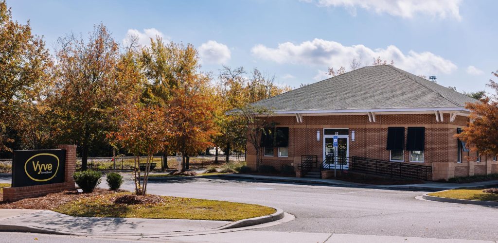 A one-story brick building with a gray roof and black shutters, surrounded by autumn trees. A sign reading “Vyve” stands near the driveway entrance. The sky is partly cloudy.