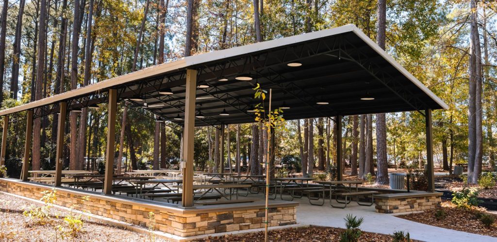 A covered picnic pavilion with multiple tables and benches is set in a wooded park area with tall trees and autumn leaves on the ground. The pavilion has a metal roof and stone accents around the base.