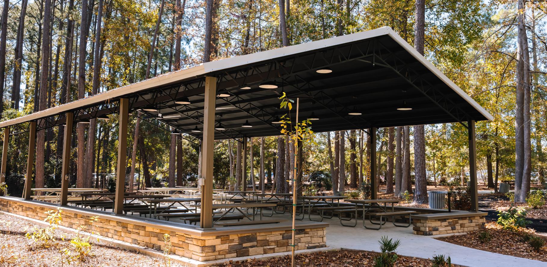 A covered picnic pavilion with multiple tables and benches is set in a wooded park area with tall trees and autumn leaves on the ground. The pavilion has a metal roof and stone accents around the base.