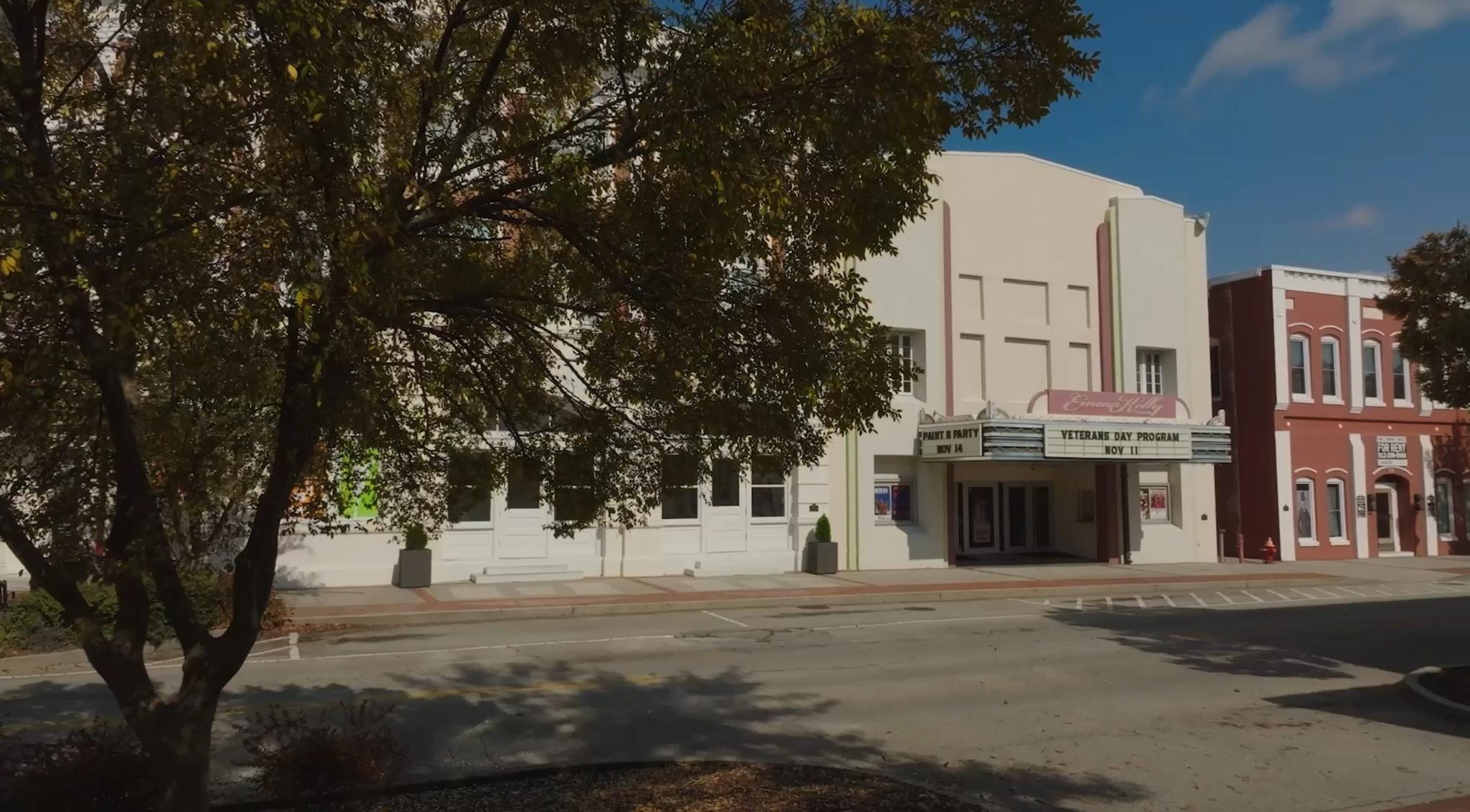 A historic theater with a retro marquee stands on a quiet street, partially shaded by a large tree. The building is cream-colored with red trim, and old-fashioned storefronts are visible to the right.