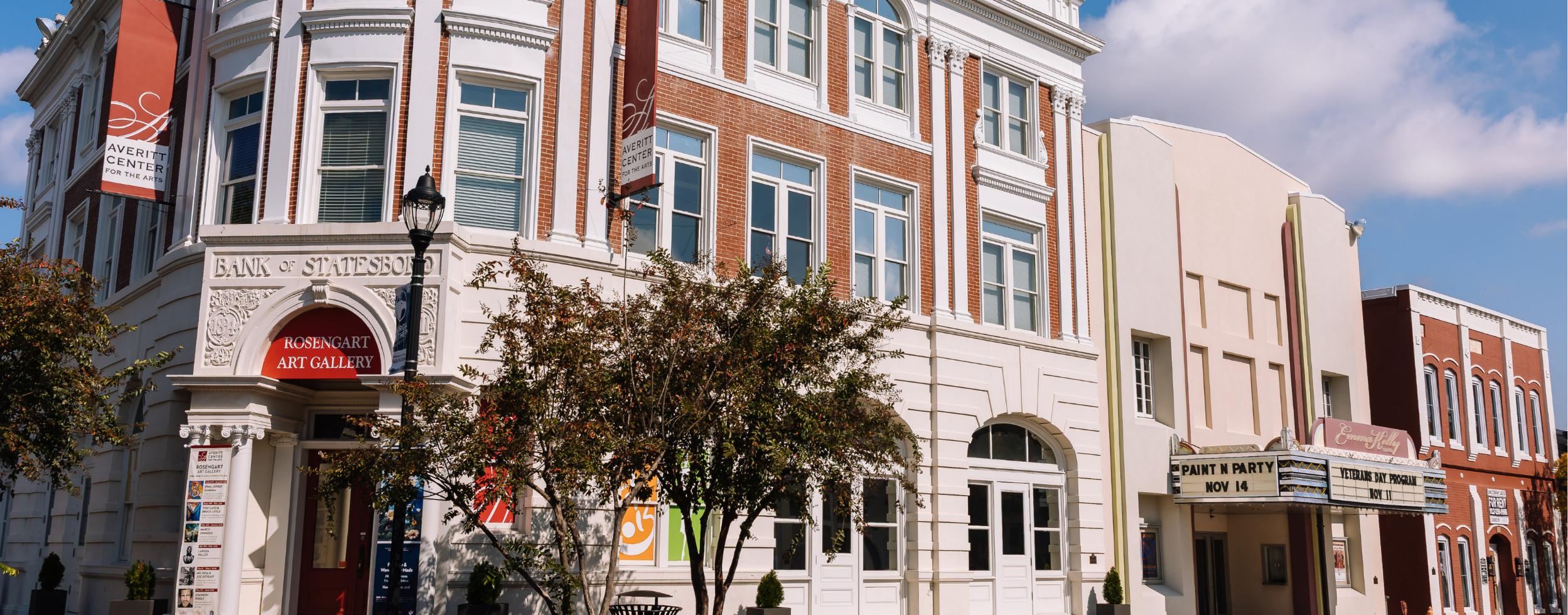 Historic brick and white building with banners for an art gallery, trees in front, and an old-fashioned movie theater marquee next door under a blue sky.