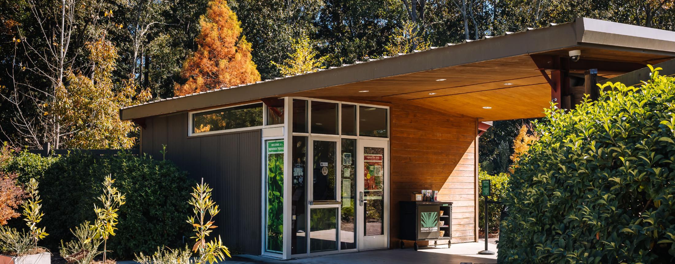 A modern building with large glass doors and a slanted roof is surrounded by lush green bushes and tall trees with autumn foliage under a clear sky.