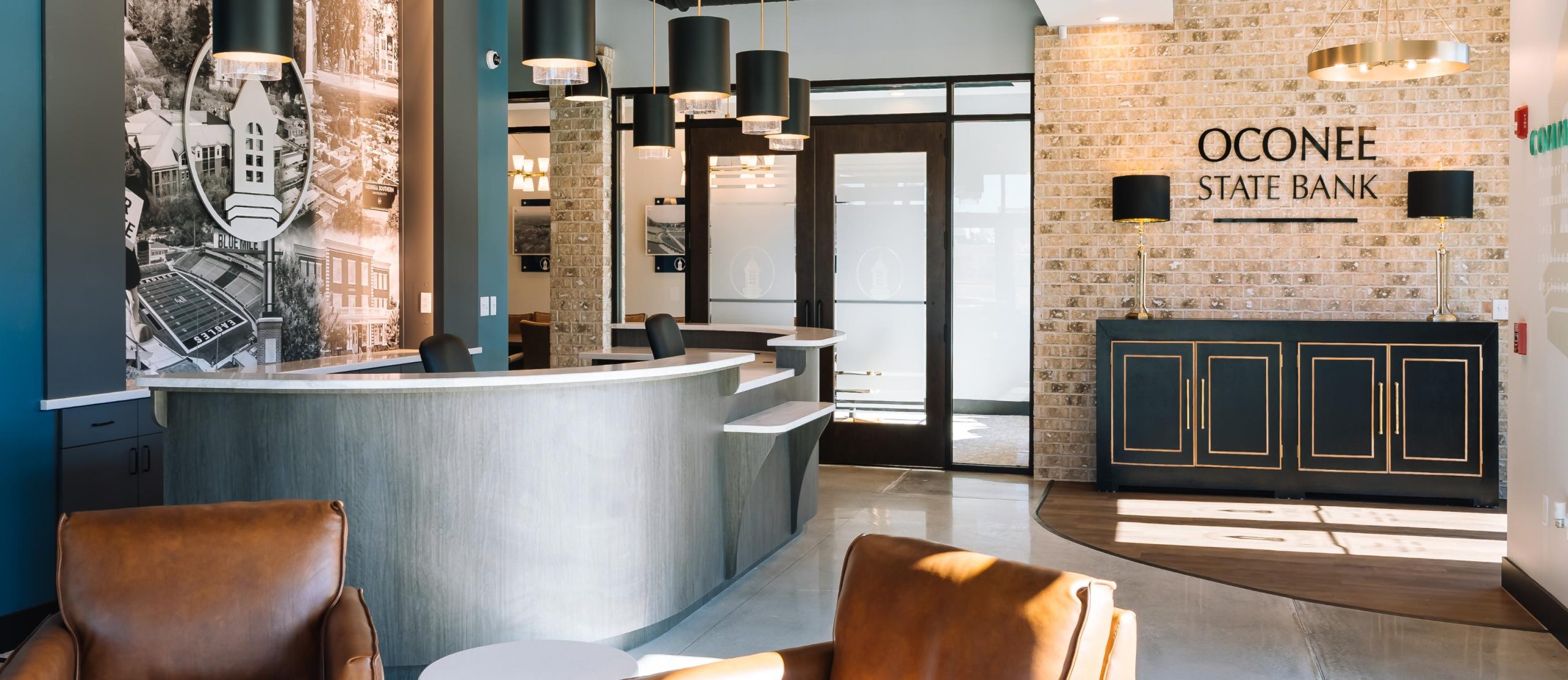 Modern bank lobby with curved reception desk, brown leather chairs, pendant lighting, large wall art, and a sign reading Oconee State Bank on a brick accent wall. Glass doors and windows let in natural light.
