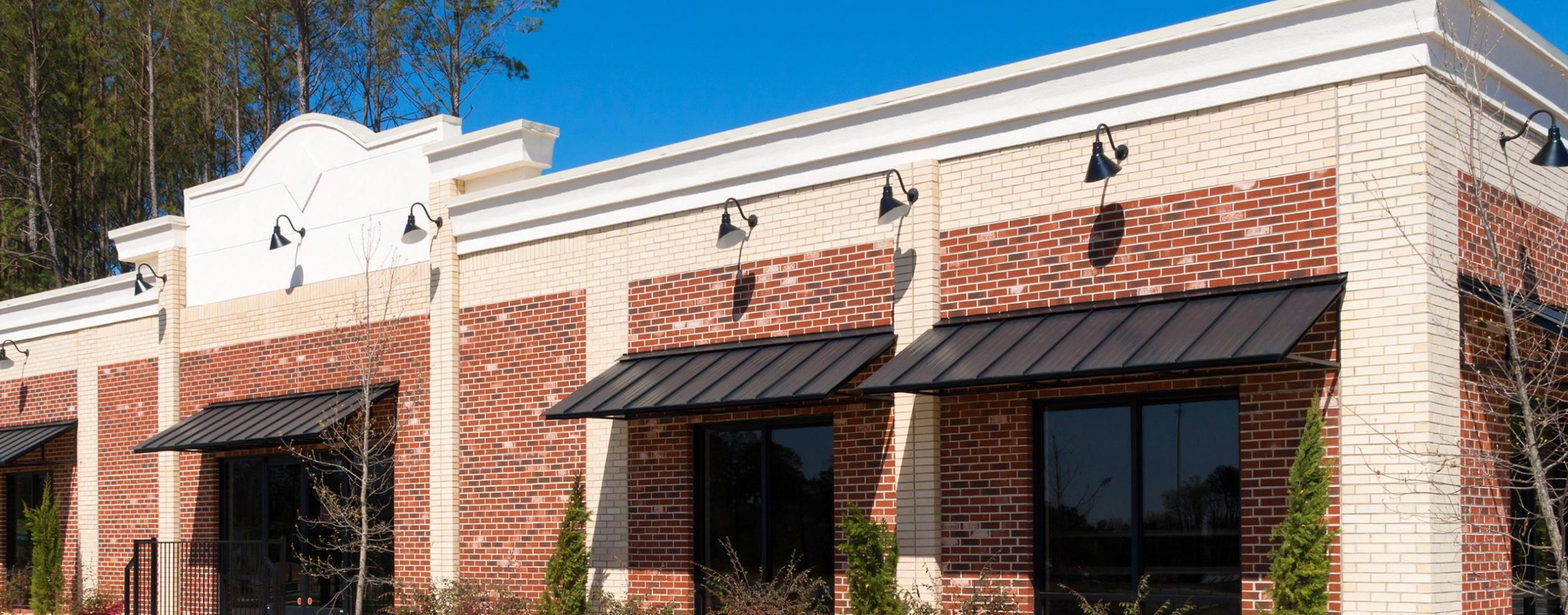 A modern, single-story commercial building with red and white brick exterior, black awnings above large windows, and outdoor wall lights, surrounded by small shrubs and trees under a clear blue sky.