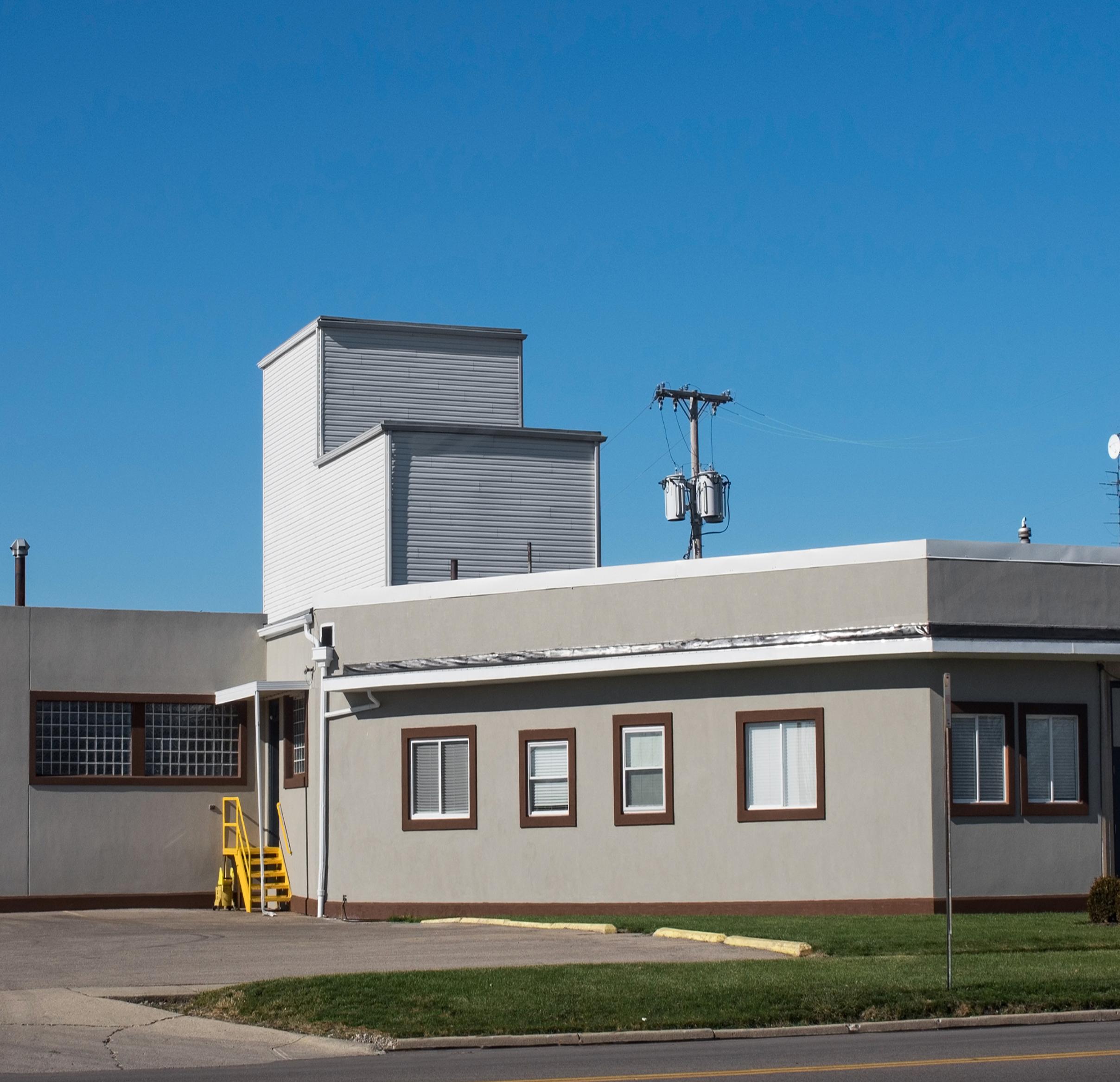 A plain, modern building with white and brown trim, small windows, and a set of yellow stairs outside. The sky is clear and blue, with some power lines visible in the background.