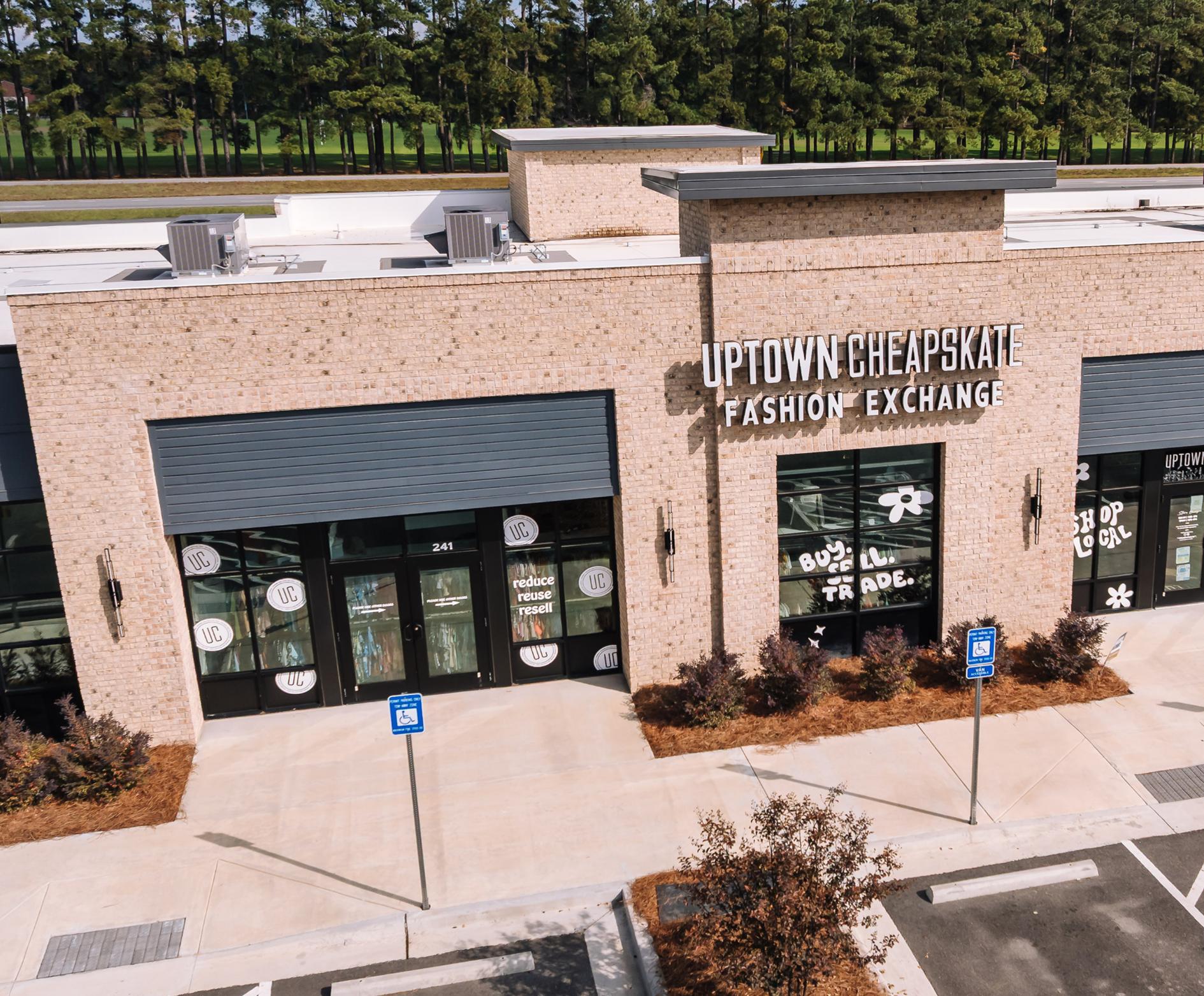 A tan brick building with a sign reading Uptown Cheapskate Fashion Exchange. Large windows display decorative designs. There are empty parking spaces and two blue accessible parking signs in front. Trees line the background.