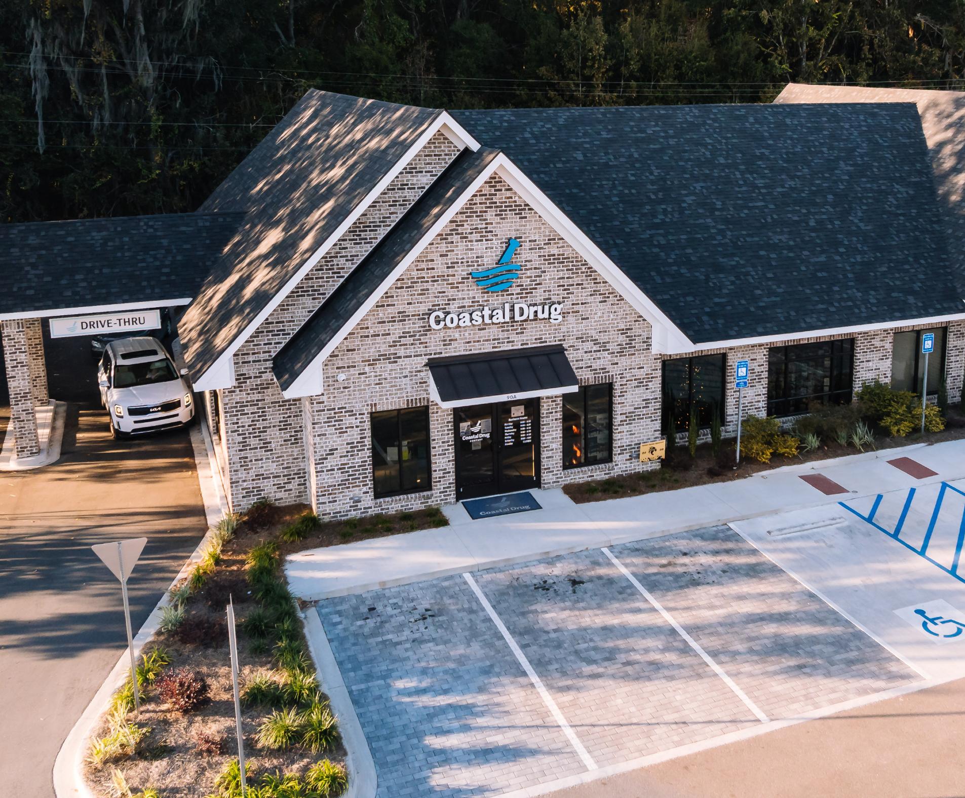 Aerial view of a brick building with a sign reading Coastal Drug above the entrance, a drive-thru lane to the left with a white car, and an adjacent parking lot with marked spaces, including accessible parking.