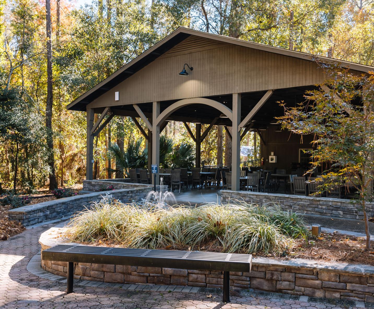 A covered outdoor pavilion with tables and chairs sits among tall trees and landscaped plants. A stone wall and a black metal bench are in the foreground, and sunlight filters through the leaves.